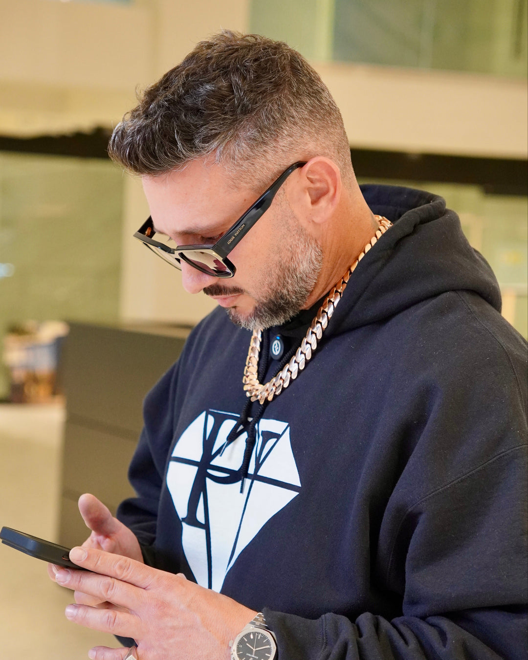 Man wearing glasses and a dark hoodie with a white logo, using a phone.
