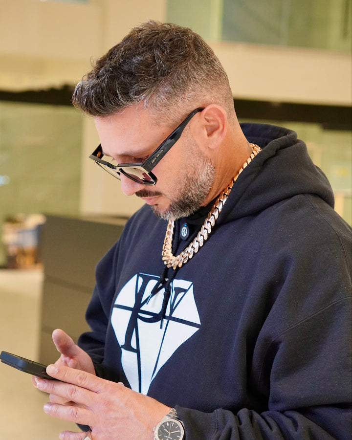 Man wearing glasses and a dark hoodie with a white logo, using a phone.